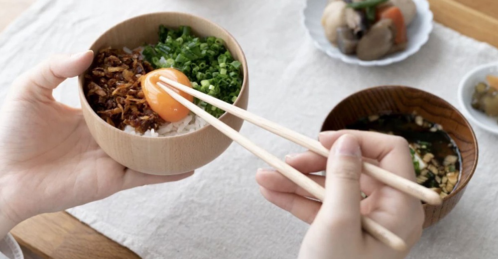 一雙淺色楓木圓筷，夾著碗中的飯、炸物、蔥花和生蛋黃，另一隻手持筷準備夾取碗裡的湯。