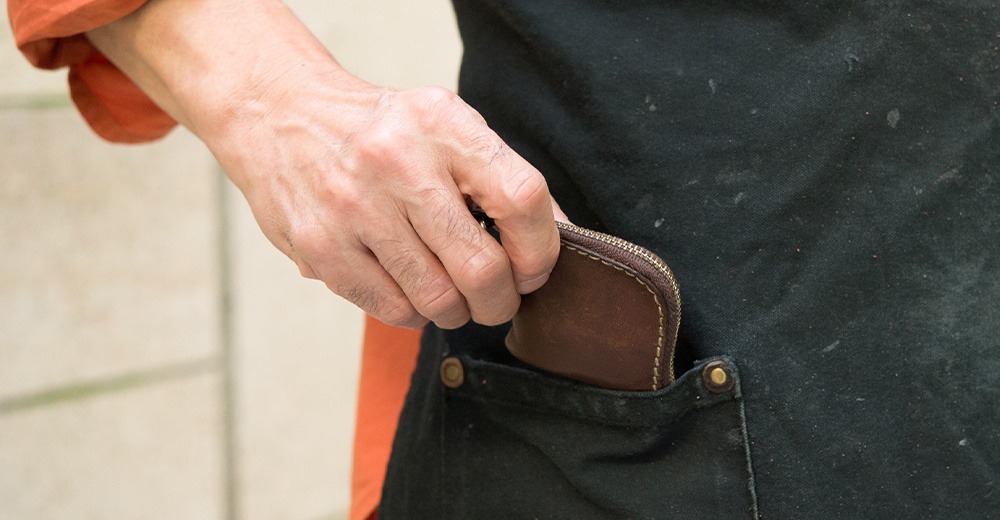 一位穿著黑色圍裙的人，正將一個精緻的深棕色日本HERZ手作皮革零錢包放入圍裙的口袋中，展現出皮革的質感與職人手工製作的細節。