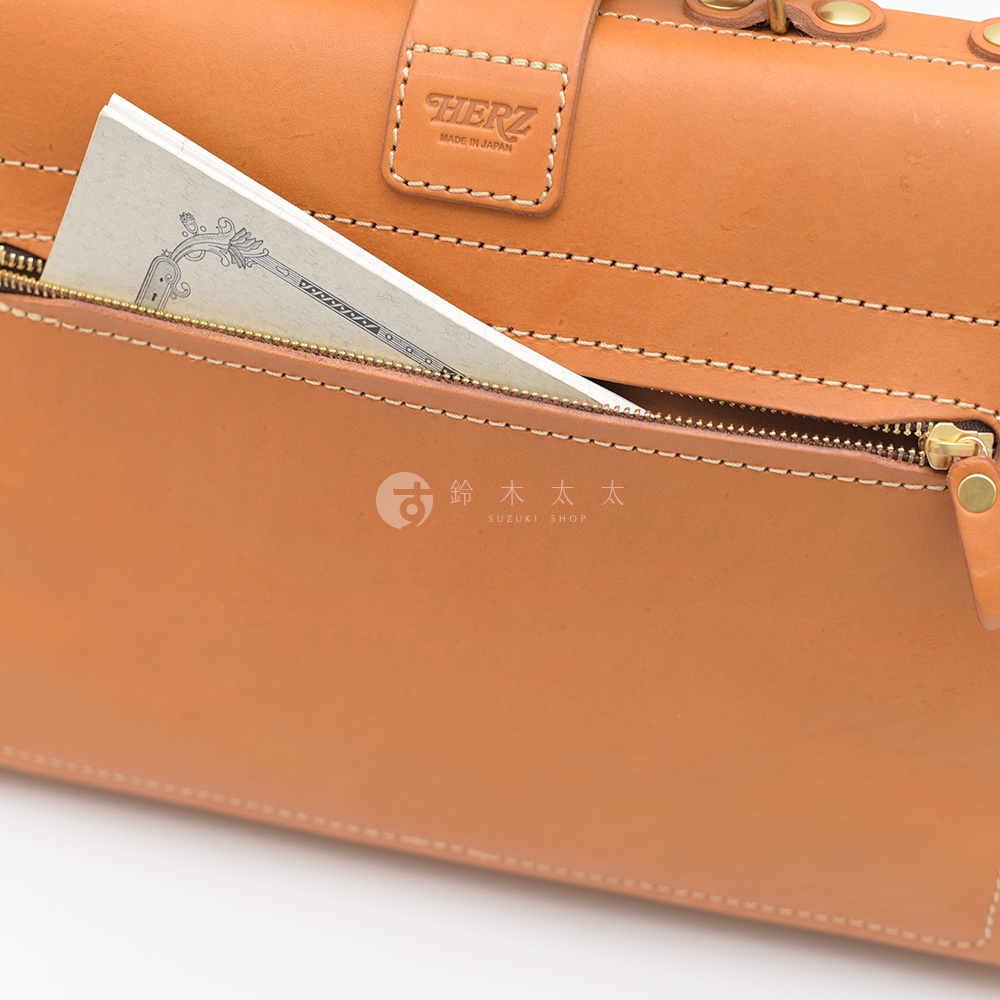職人手作皮革軟式杜勒斯包，淺棕色皮革，拉鍊口袋露出部分紙張，包身有品牌燙金標誌。