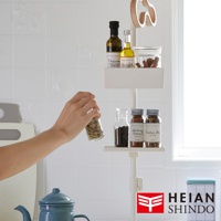 白色伸縮桿上層架著橄欖油瓶、調味料罐及肉桂棒，下方架著黑胡椒粒和混合香料罐，一隻手正將香料罐放到架上，背景為白色磁磚牆和窗戶。