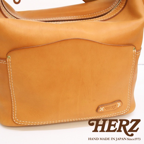 棕褐色厚實皮革單肩波士頓包，正面有弧形翻蓋口袋，白色車線縫製，右下方有品牌壓紋標誌。