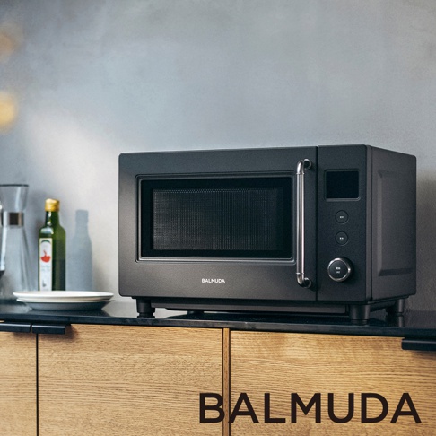一台放在木紋櫥櫃檯面上的黑色 BALMUDA 微波爐，外觀呈現簡約的金屬質感，右側設有旋鈕與控制面板，左側旁擺放著一個空玻璃水瓶及一疊白色餐盤，背景為灰色的牆面。