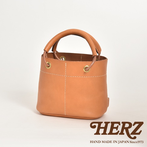 職人手作皮革加厚提把大開口午餐袋，採用柔軟的駝色皮革製成，設有加厚的圓形提把，透過縫線勾勒出立體感。袋口配有金屬孔和皮革抽繩，設計簡潔實用。