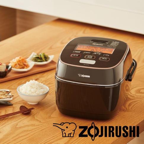 深棕色Zojirushi電子鍋，帶有銀色飾邊和觸控面板，面板上有日文按鈕和顯示螢幕。旁邊擺放著一碗白米飯、幾碟日式小菜和一雙筷子。