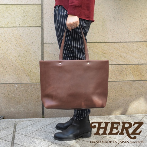 一款棕色皮革托特包，配有兩條皮革提把和兩個金屬鉚釘固定。包款設計簡潔，容量寬敞，適合日常使用。