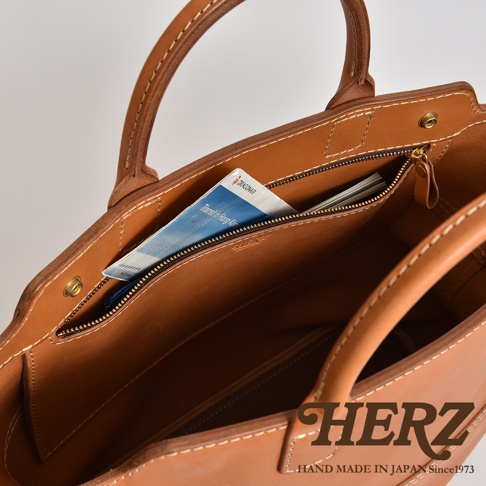 淺棕色皮革托特包，內有拉鍊口袋，可見一本藍色封面的雜誌，包包縫線精緻，展現皮革質感。