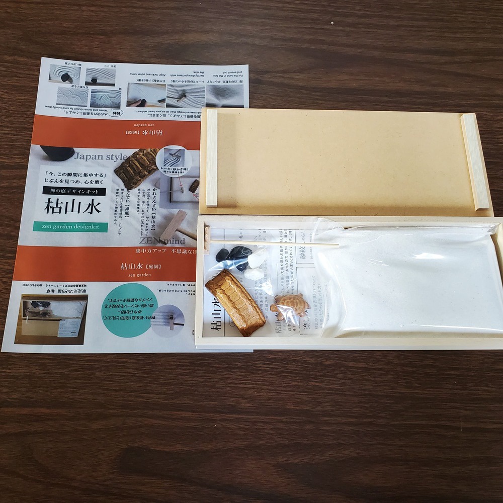 日式枯山水模型庭園設計套組，包含木質外盒、說明書、白色細砂、黑白石子、木製耙子、以及裝飾用的木製建築模型與烏龜模型。