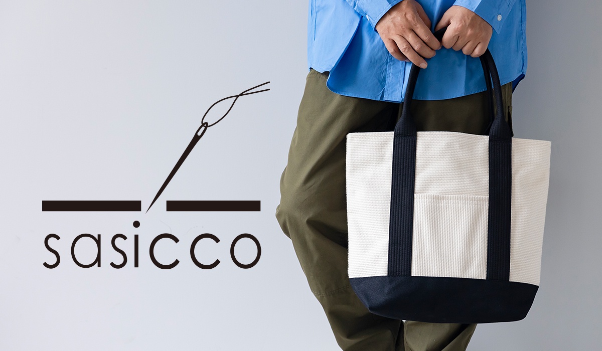 一個身穿藍色襯衫和卡其色褲子的人，右手提著一個米白色和海軍藍的帆布托特包，左側有一個帶有針線圖案和「sasicco」字樣的品牌標誌。