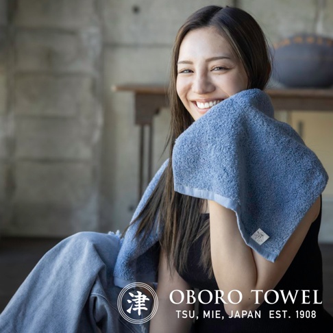 一位女性手持一條藍色毛巾，毛巾材質柔軟，邊緣有細緻縫線，毛巾上印有品牌標誌。