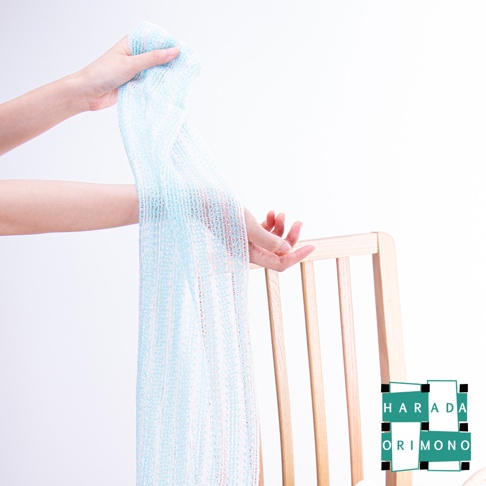 一雙手正拉著一條淡藍色的泡泡洗澡巾，這條洗澡巾由細密的網狀材質製成，帶有細小的顆粒，呈現出立體紋路。背景為白色，右側有木質椅子的局部。