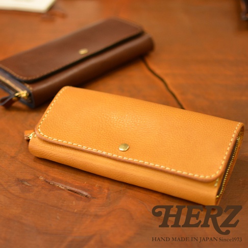 兩個職人手作皮革長夾，一個為芥末黃色，另一個為深棕色，皆為寬口設計，表面有細緻紋理，並以白色車縫線勾勒邊緣，搭配金色壓扣與拉鍊。