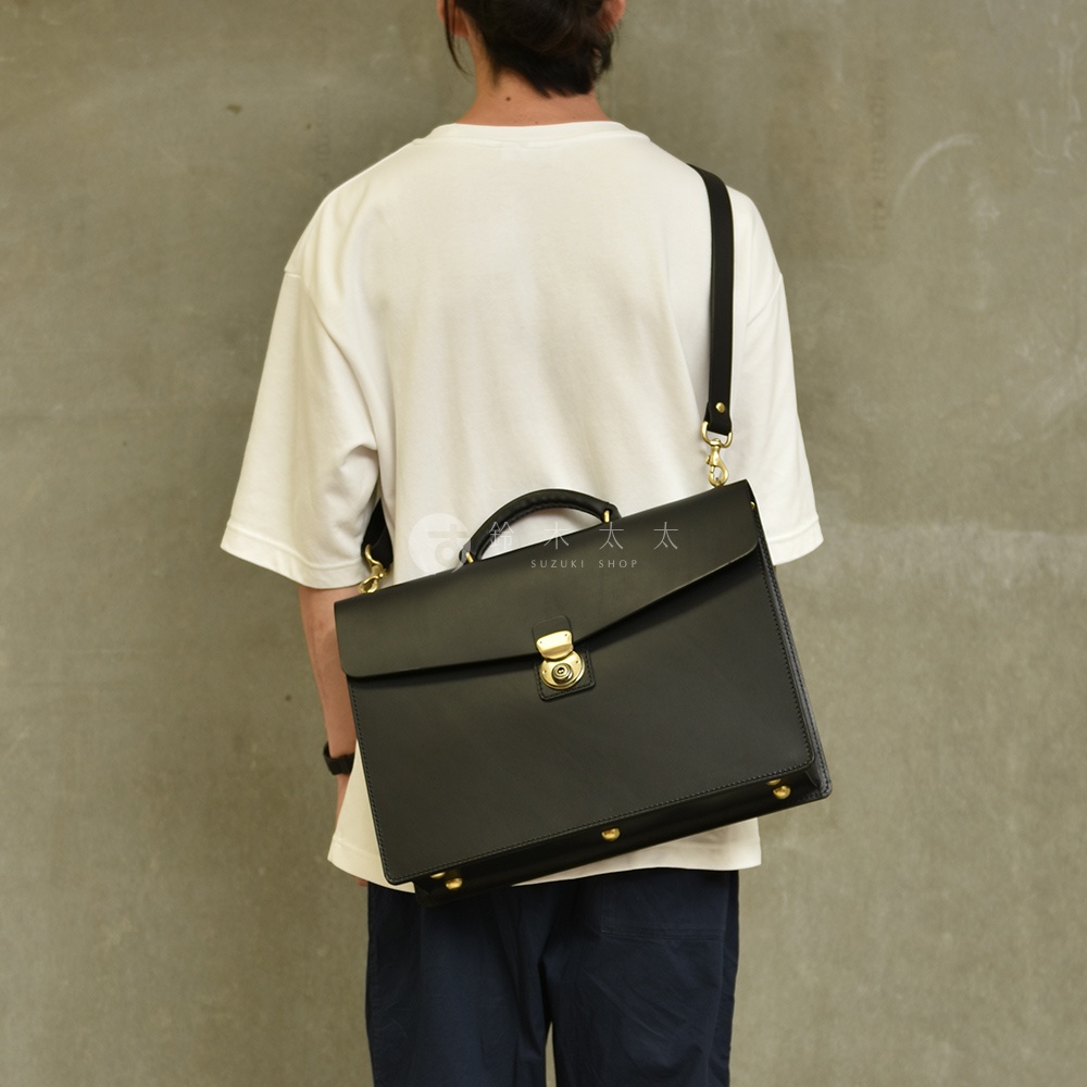 黑色皮革兩用商務公事包，配有金色旋轉鎖扣、頂部提把及可拆卸肩帶，包身底部有金色金屬釘裝飾。