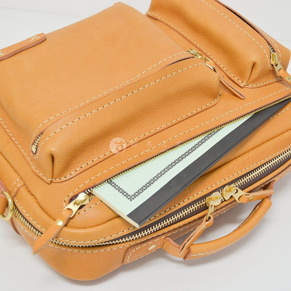 職人手作的淺棕色皮革兩用包，展示了包身多個拉鍊口袋和手提握把，其中一個拉鍊口袋內有綠色筆記本。