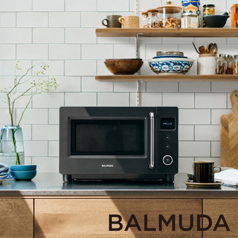廚房檯面上放置著一台黑色 BALMUDA 微波爐，其簡約的現代風格與旁邊的青色玻璃花瓶、木質層架上的器皿和調味瓶形成鮮明對比。微波爐數位面板顯示著 HELLO 字樣，整體畫面呈現出充滿生活感的家居氛圍。