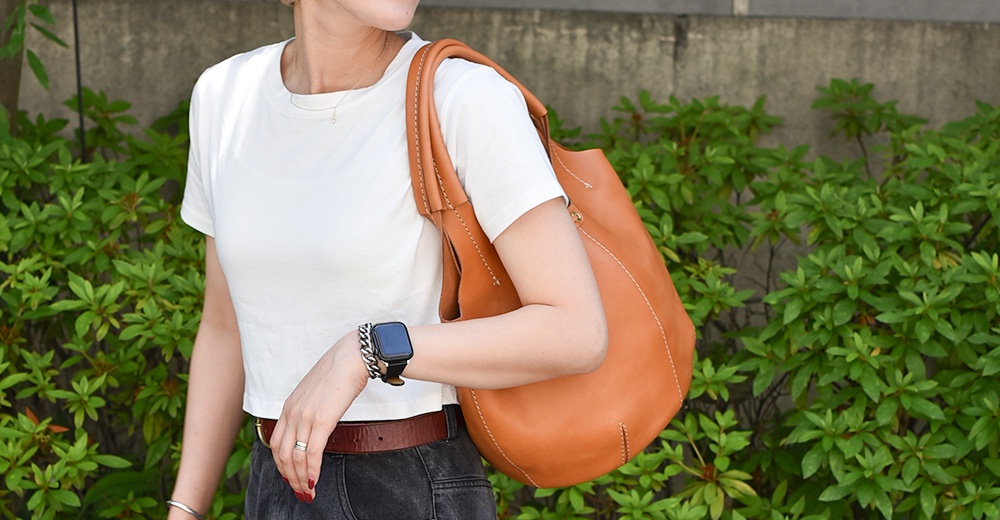 駝色皮革球形肩掛托特包，配有寬版皮革肩帶，包身縫線明顯，由植物背景襯托。