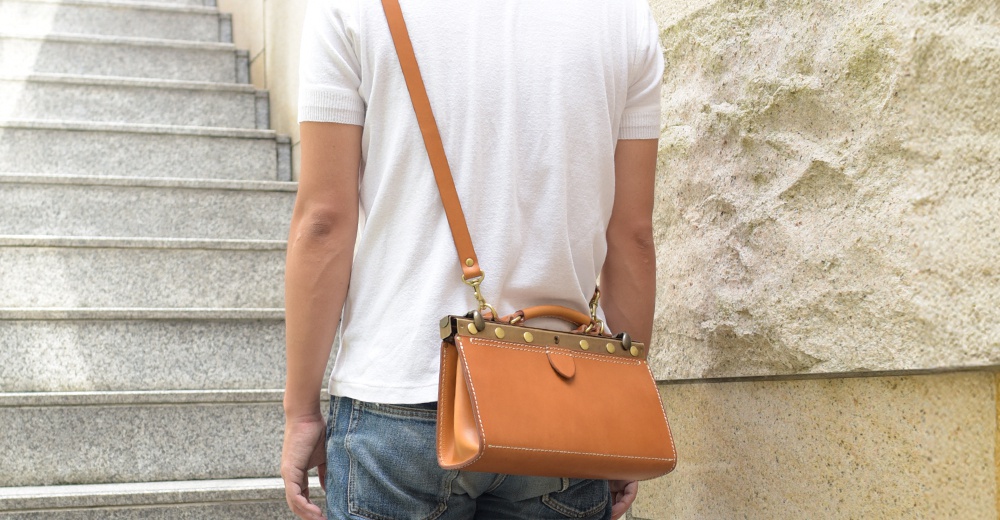 一名男子斜挎著一個焦糖色皮革復古口金兩用手提包，包包配有可拆卸的長肩帶和頂部提把，包邊有縫線裝飾，並有金屬口金和釘釦。