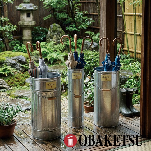 OBAKETSU 手工製鍍鋅鋼傘桶，擺放於日式庭園木製地板上，桶身內插著多把木柄雨傘，雨中景緻。