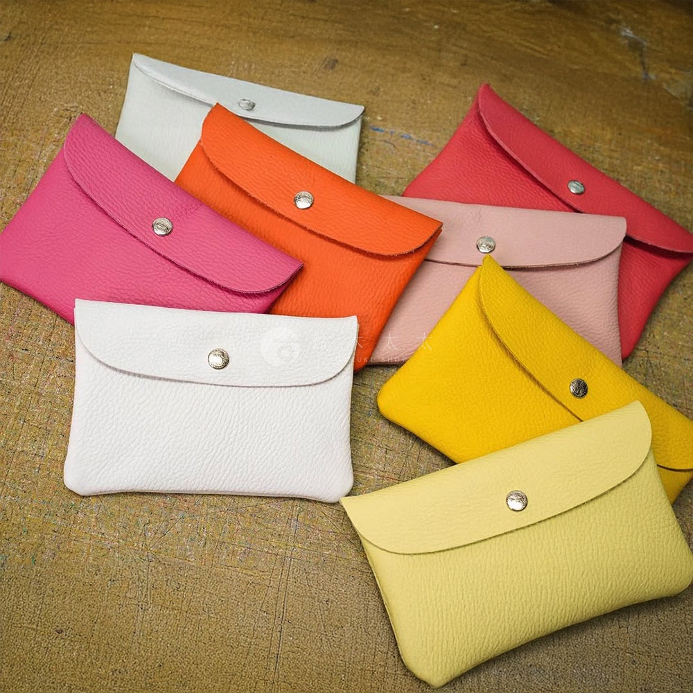 多個京都職人手作多功能收納包，呈扇形散開。包款為荔枝紋皮革材質，顏色包含粉紅、橘紅、淺粉、亮黃、淺黃、淺灰及白色，皆有銀色按扣。