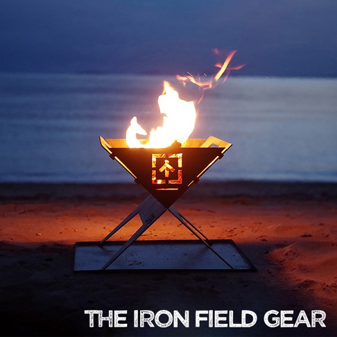 TAKI BEITA L 尺寸焚火台爐架套裝，黑色金屬材質，中央有方形箭頭鏤空設計，下方為 X 型支架，置於沙灘上，火焰燃燒中。