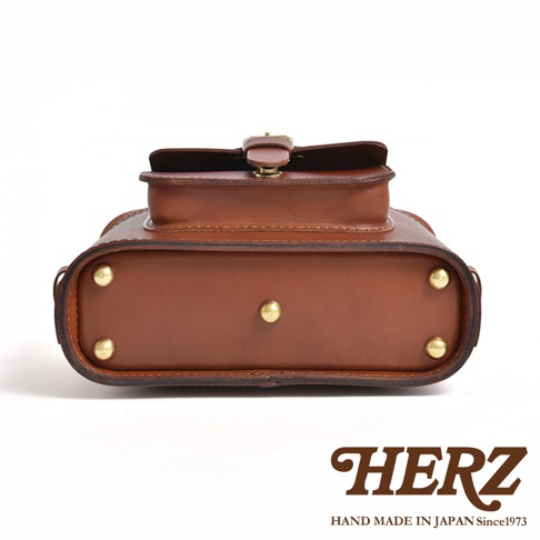 HERZ 品牌 logo 與「HAND MADE IN JAPAN Since1973」字樣，下方為棕色皮革相機包的底部視角，飾有金屬釘。