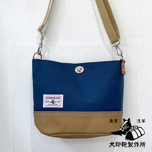 犬印純綿帆布雙色迷你側背包，深藍色與卡其色拼接，配有可調式卡其色帆布背帶，銀色金屬開關和掛鉤，正面有品牌標籤和圖案。