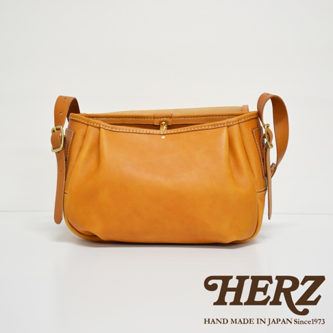 職人手作皮革復古翻蓋郵差包，採用溫暖的焦糖棕色皮革，搭配可調節的皮質背帶與銅質金屬扣。包身設計簡約，有著自然的皮革光澤與精緻的縫線細節。