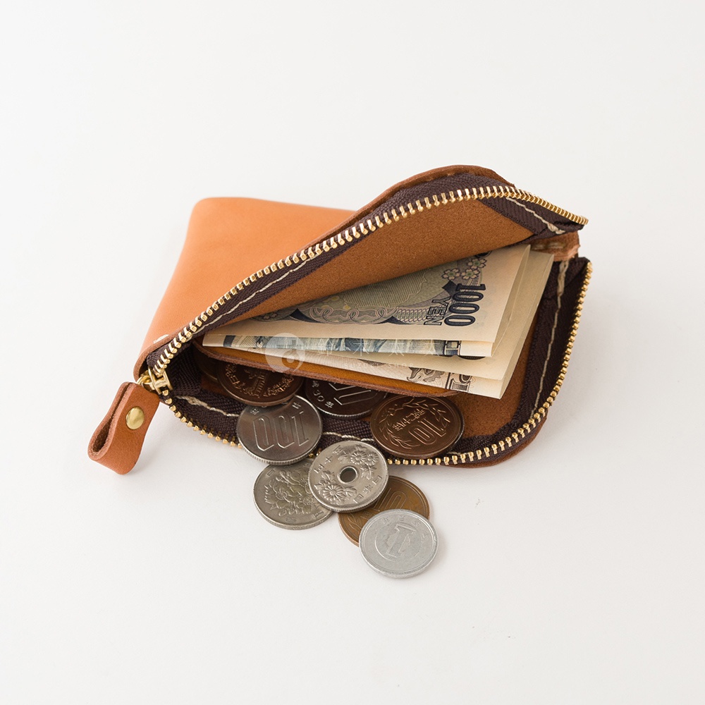 一個開口的棕色皮革迷你零錢包放置在白色背景上，錢包內部裝有日本千元紙鈔，袋口處散落著多枚不同面額的日本硬幣，展現簡約實用的收納功能。