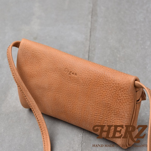 駝色紋理皮革製成的斜背包，上面壓印著Organ字樣，底部右側有HERZ字樣及HAND MADE IN JAPAN SINCE 1973的字樣。