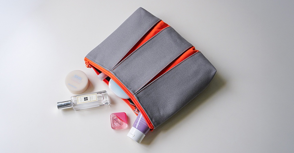 一個灰色帆布材質的長方形收納包，內部飾有亮橘色內襯，開口處露出化妝品與保養品。收納包周圍散落著香水瓶、圓形粉餅盒、小巧的粉紅色藥水瓶以及一支紫色軟管裝乳液，整體呈現出簡潔、現代的收納美學。