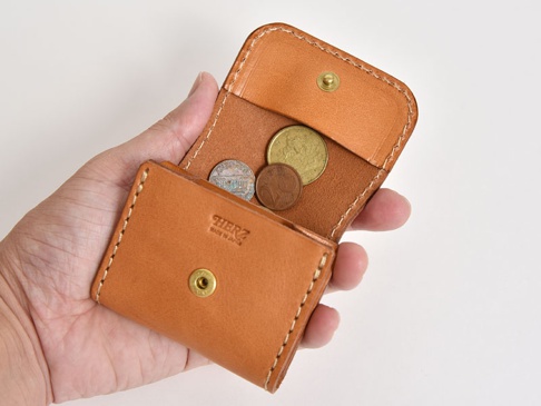 一只手拿著一個淺棕色皮革雙折收納零錢包，包內裝有數枚硬幣。零錢包有縫線細節和金屬按扣，表面印有「HERZ MADE IN JAPAN」字樣。