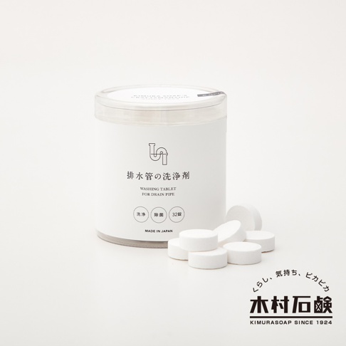 白色圓形塑膠罐裝著排水管清潔錠，罐身印有黑色圖案和「排水管の洗浄剤」日文及「WASHING TABLET FOR DRAIN PIPE」英文。罐旁散落幾顆白色圓柱形清潔錠。右下方有日文及英文品牌標誌。