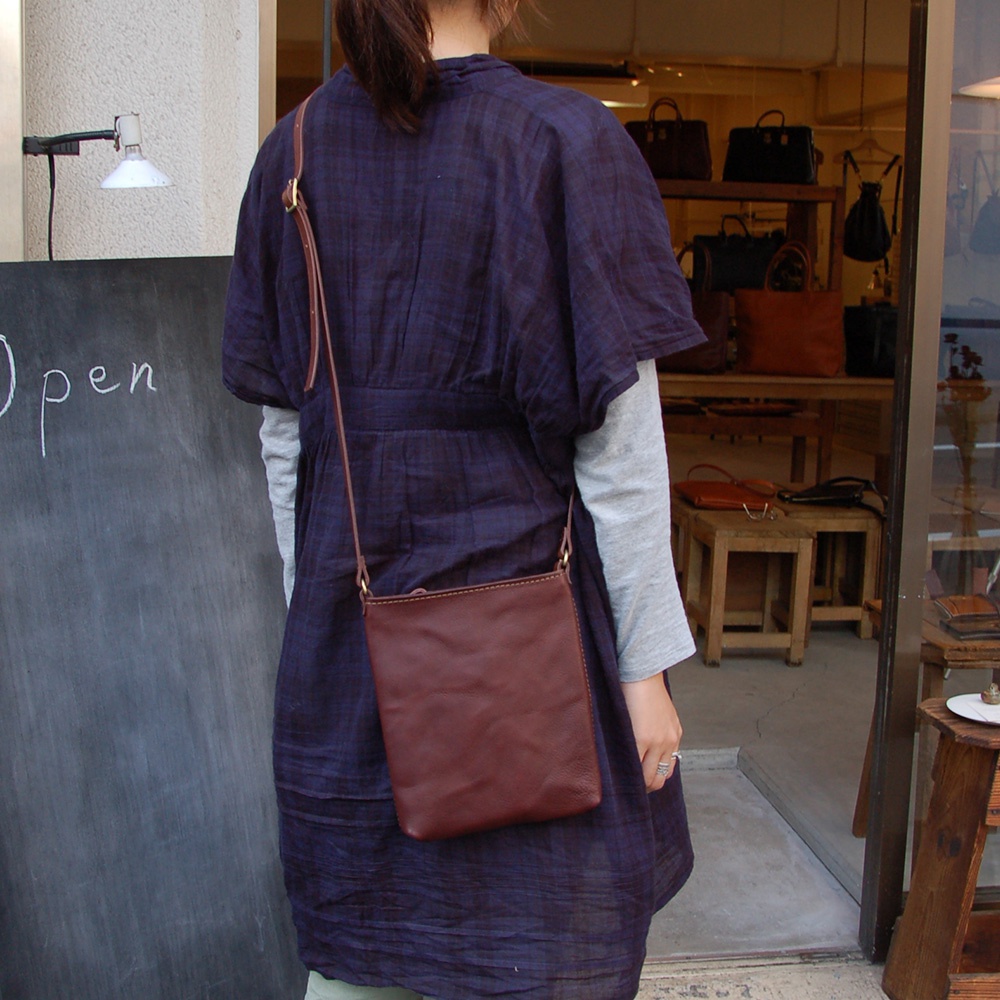 棕色皮革製成的長方形肩背包，以細長肩帶斜背於身穿深藍色格紋洋裝的模特兒身上，包款線條簡潔。