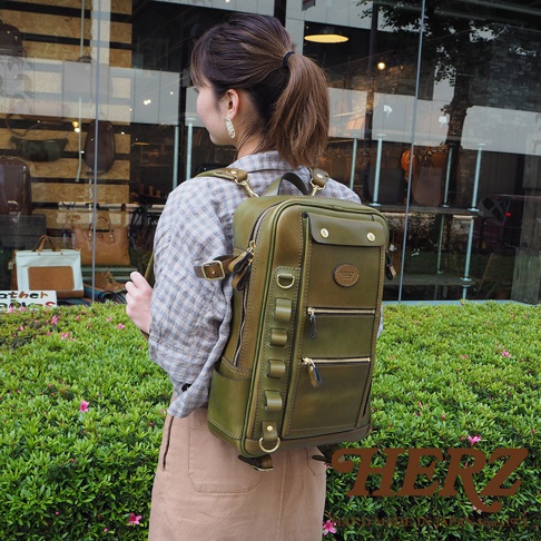 一名女性身背綠色皮革兩用多功能後背包，背包為長方形直立設計，正面有品牌標誌、多個拉鍊口袋和可調節的皮帶裝飾。