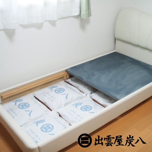 白色床架內，整齊擺放著六個印有藍色日文「炭八」字樣的白色包裝袋，部分包裝袋的角落可見「高調濕 NET 12L」等字樣。床架上方覆蓋著深灰色布墊，床頭為白色皮革材質。