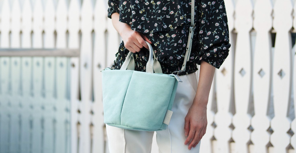 一個穿著碎花襯衫和白色長褲的人，手裡拿著一個淺綠色的斜背包，背景是白色欄杆。
