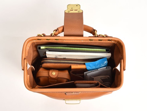 職人手作皮革兩用迷你口金單肩包 (CW-7) 頂部視角，展示包內裝有筆記本、錢包、小皮件及藍色包裝物，整體為淺棕色皮革材質，配有金色金屬口金。