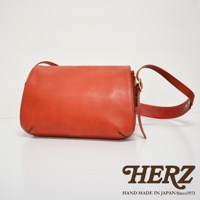 一個橘紅色皮革復古翻蓋郵差包，有可調節的皮質肩帶，包身縫線清晰可見。