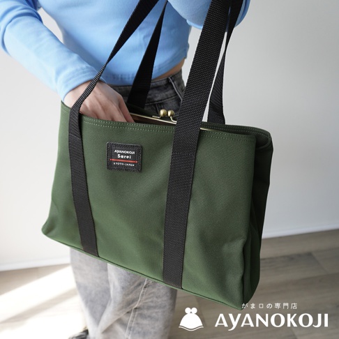 A green AYANOKOJI Sarei Kyoto-Japan tote bag with black straps is held by a person wearing a light blue shirt and gray jeans.