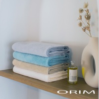 疊放的今治毛巾，由上至下顏色分別為淺藍、藍綠、米黃、白色，質地看起來柔軟吸水。旁邊有一個裝有透明液體的玻璃瓶和一個白色陶瓷花瓶，裡面插著乾燥的植物。