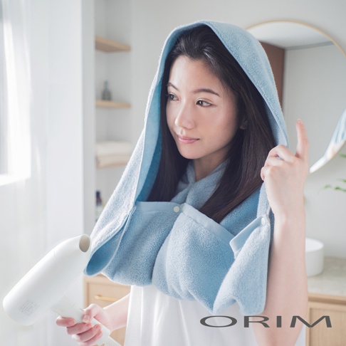 一位女性使用藍色吸水毛巾頭巾，頭巾有扣子固定，肩部包裹著毛巾，她正用吹風機吹乾頭髮。