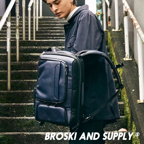 深藍色防水真皮革兩用手提後背包，有前置拉鍊口袋與下方翻蓋口袋，搭配黑色背帶，由男性背負於樓梯間。