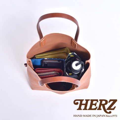 一個日本HERZ手工製作的棕色皮革托特包，俯視角度展示了包內收納空間，包中裝有綠色皮夾、黑色手機、紅色拉鍊錢包及一把黑底帶有圓點圖案的折疊傘，圖片右下方印有HERZ品牌標誌及HAND MADE IN JAPAN Since1973字樣。