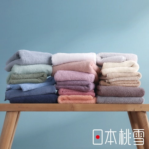 一張放置在木製長凳上的日本桃雪精梳棉飯店毛巾堆疊圖，毛巾呈現多種柔和的莫蘭迪色系，包括藍色、綠色、白色、粉色及大地色等，畫面右下角帶有日本桃雪品牌標誌與紅色圓點設計，背景為簡約的淺藍色。