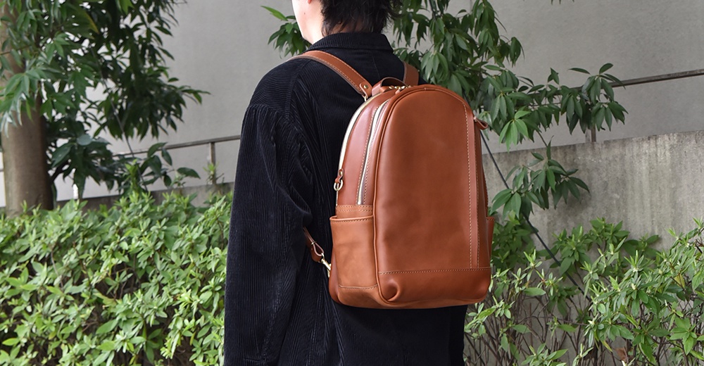 一件棕色皮革多功能後背包，背包正面有拉鍊開合和側邊口袋，由一位身穿深色燈芯絨外套的人背著，背景為綠色植物和水泥牆。
