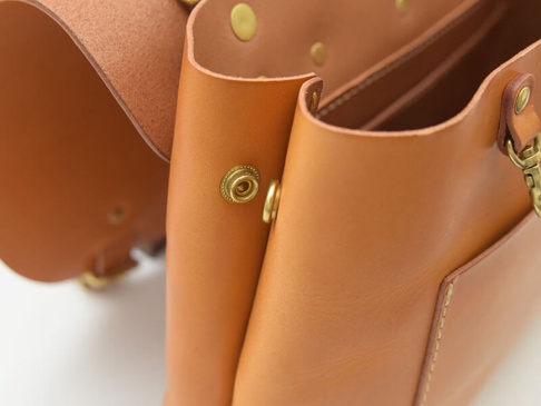 職人手作的棕色皮革雙扣三用後背包，特寫展示其細緻的皮革紋理、黃銅色雙扣和金屬鉤環，以及乾淨的車縫線細節。