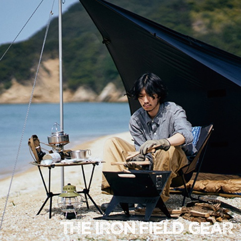 TAKI BEITA焚火台爐架套裝 L，黑色金屬材質，包含一個折疊式焚火台，旁邊是露營桌、野營燈和鍋具，一位男性正在準備木柴。