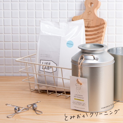 無添加洗衣粉補充包，白色紙袋包裝，附有日文說明及圓形藍色「ORIGINAL」標籤。旁邊為銀色金屬儲物罐，掛有附有衣物圖案及日文「LAUNDRY DETERGENT PLUS」字樣的吊牌。另有兩個金屬夾和一個木製砧板。