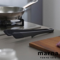 marna TOKYO 1872 標誌旁為一組灰色食物夾，後方為廚房爐具上的不鏽鋼鍋。