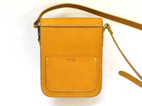 黃褐色皮革製成的直式單肩包，有皮扣和外部口袋，縫線明顯，附有可調整的長肩帶。