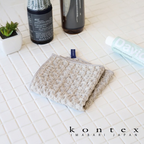 淺褐色棉麻混紡方巾，帶有鬆餅紋理，上面有藍色小標籤。背景為白色格子瓷磚，旁邊有牙膏和兩瓶洗護用品。